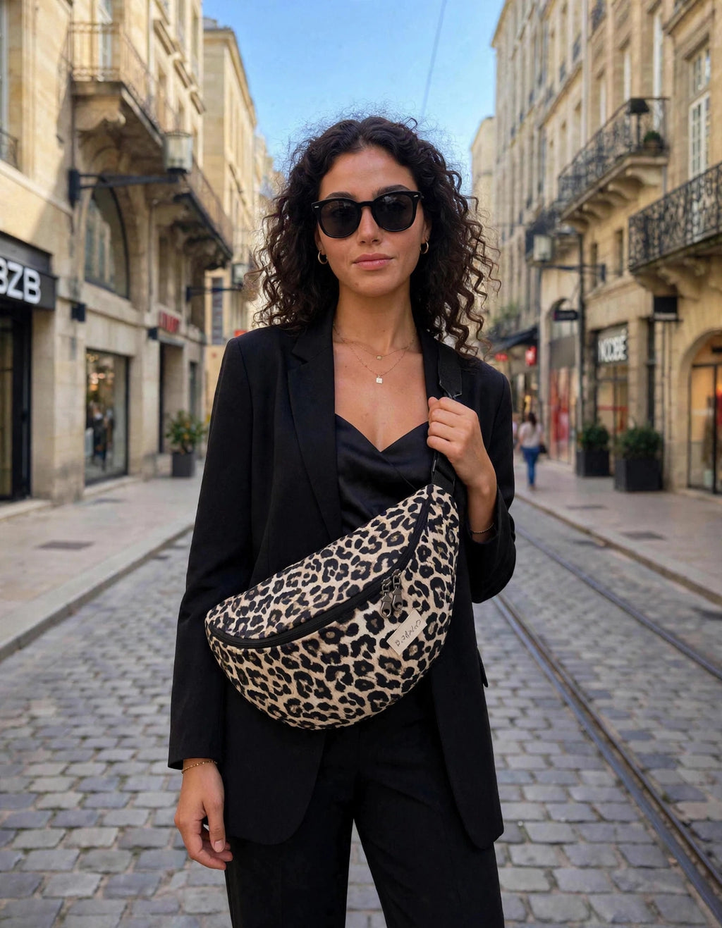 Femme portant un sac banane XL léopard Piplette & Co en bandoulière sur blazer noir, dans une rue pavée du centre de Bordeaux
